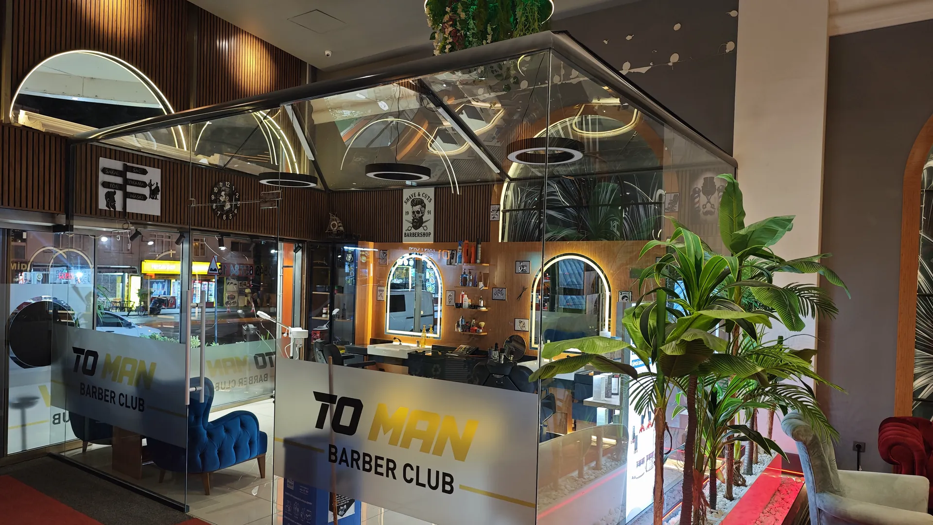 TO MAN Barber Club salon girişi, altın sarısı logo ve yeşilliklerle çevrili cam kabin